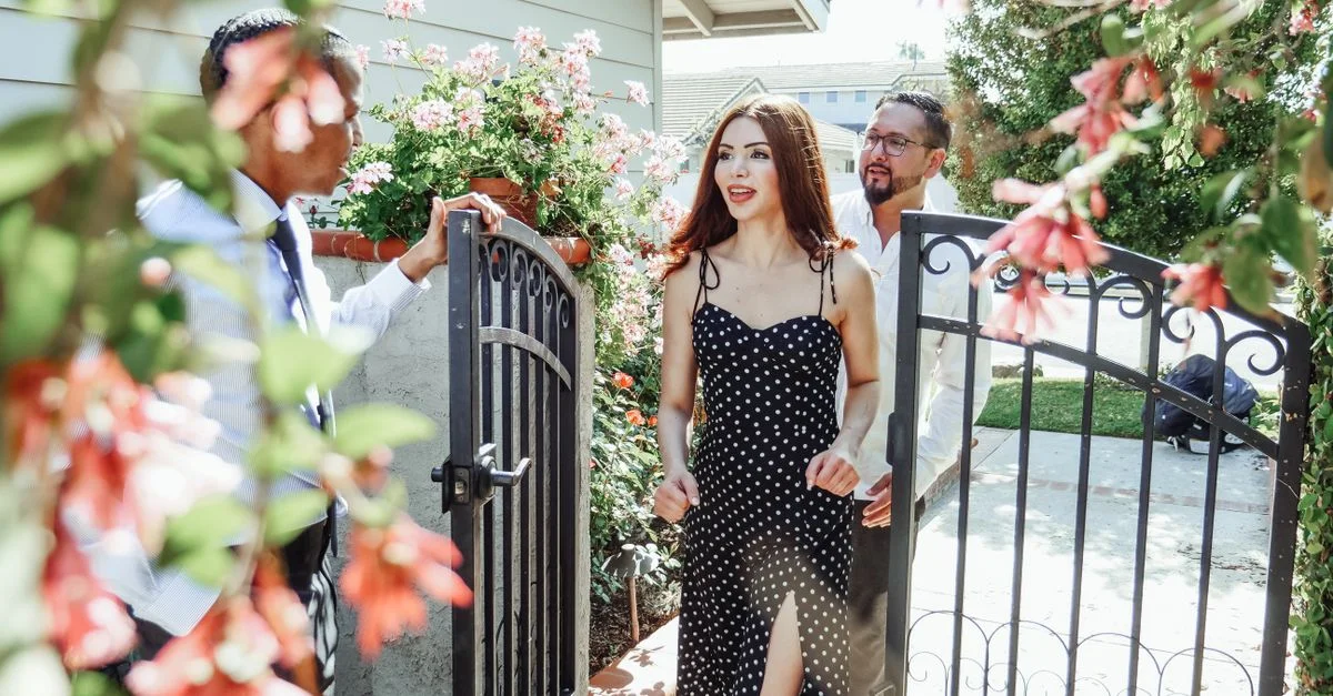 Un couple est accueilli par un agent immobilier à l'entrée d'une nouvelle maison, entouré de fleurs.