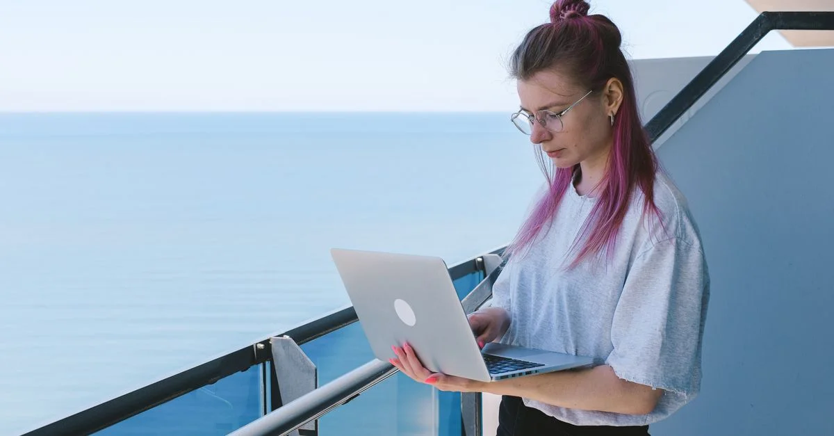 Une jeune femme travaille à distance sur son ordinateur portable au bord de la mer par une journée ensoleillée, incarnant le style de vie nomade numérique.