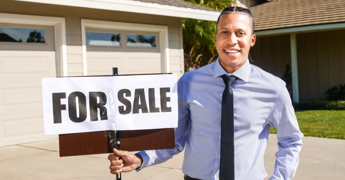 Agent immobilier souriant avec un panneau à vendre devant une maison.