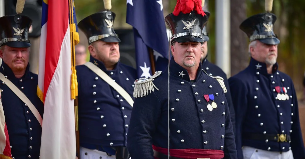 Des hommes en uniforme dans une parade militaire tenant des drapeaux et portant des médailles lors d'une cérémonie en plein jour.