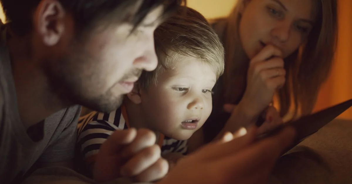 Une famille de trois personnes, composée des parents et d'un enfant, regarde attentivement une tablette numérique ensemble à l'intérieur.