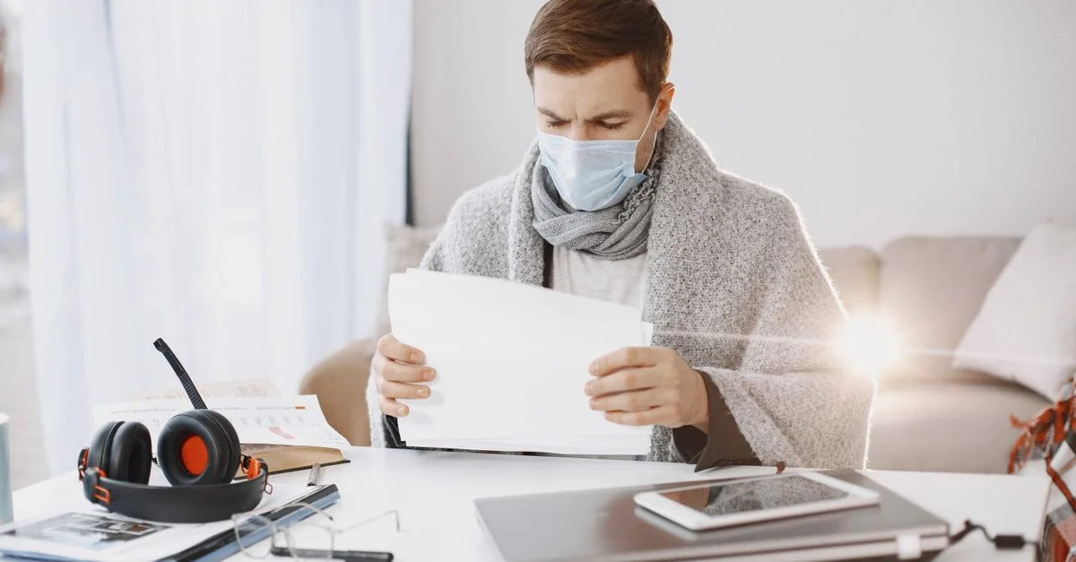 Un homme portant un masque de protection et une écharpe travaille depuis chez lui, entouré de fournitures de bureau par une journée ensoleillée.