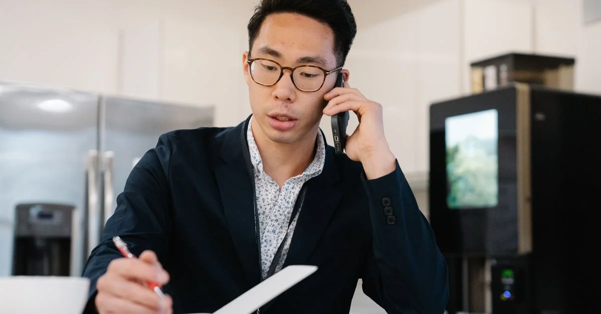 Homme asiatique en veste de costume au téléphone, prenant des notes dans un bureau.
