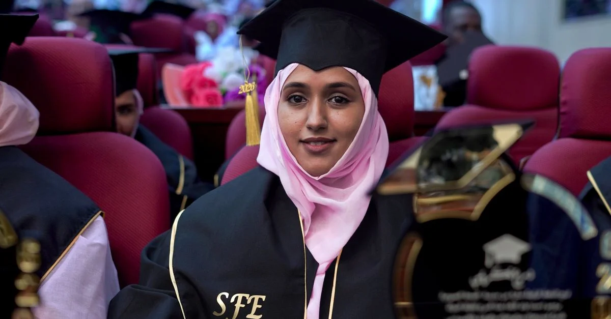 Une jeune femme portant une toque et une robe de remise de diplôme est assise dans une salle de cérémonie avec ses camarades.