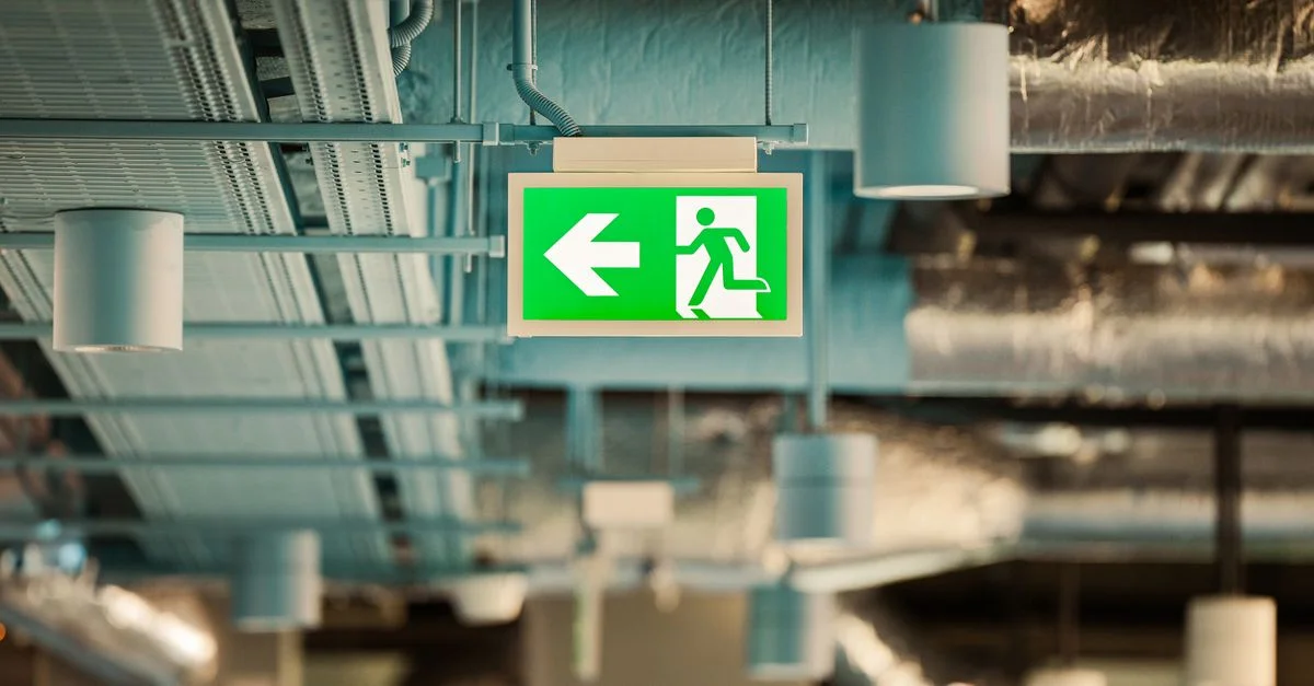 Panneau vert de sortie d'urgence suspendu à un plafond industriel, indiquant la direction vers la sécurité.
