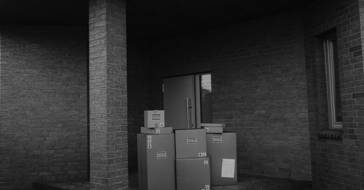 Photo en niveaux de gris montrant des colis empilés près de la porte d'un bâtiment, évoquant des thèmes de livraison moderne.