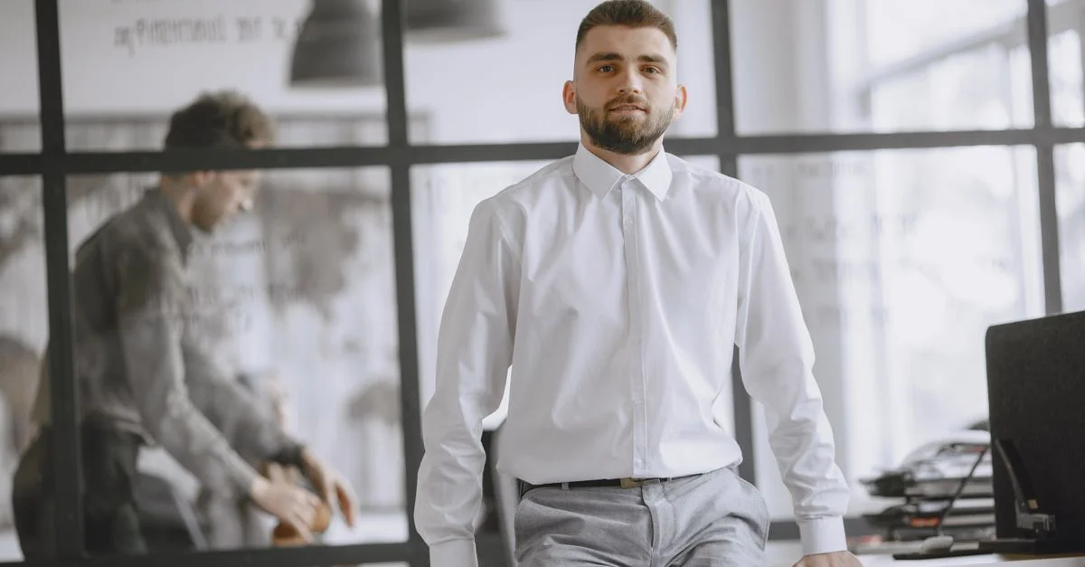 Homme confiant en chemise blanche s'appuyant sur un bureau dans un bureau moderne avec des cloisons en verre.