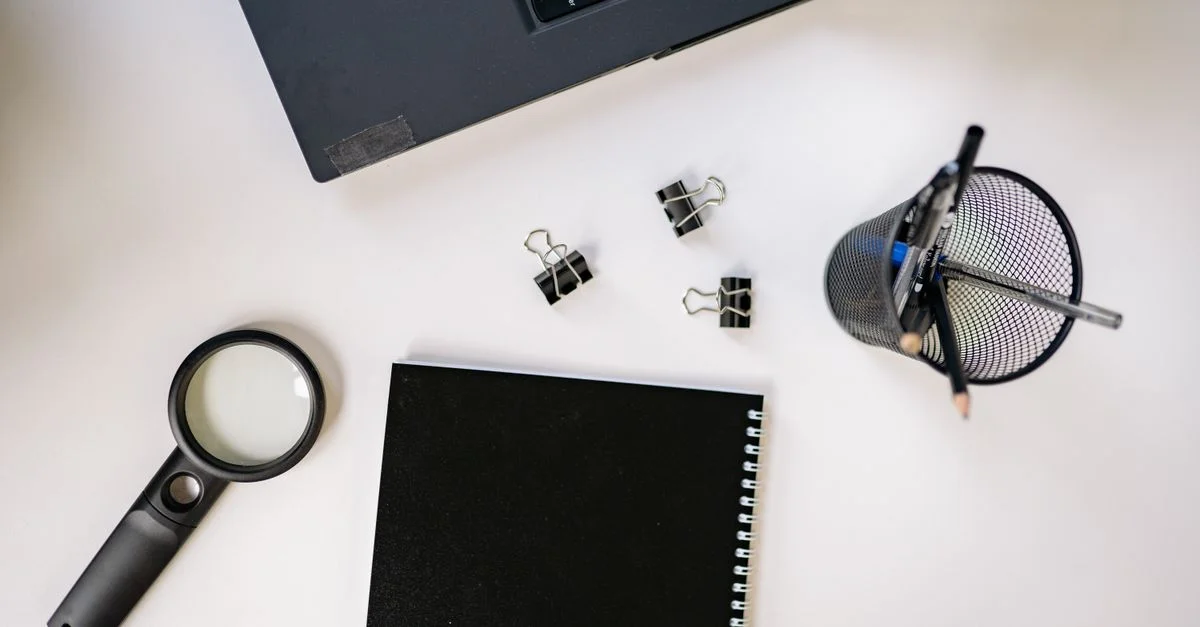 Vue de dessus d'un espace de travail avec un ordinateur portable, un carnet noir, une loupe et des fournitures de bureau sur une surface blanche.
