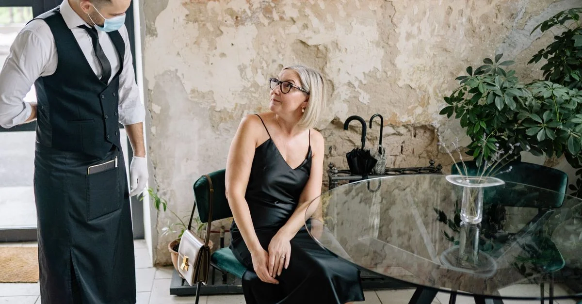 Une femme élégante en robe noire interagit avec un serveur portant un masque dans un café tendance.