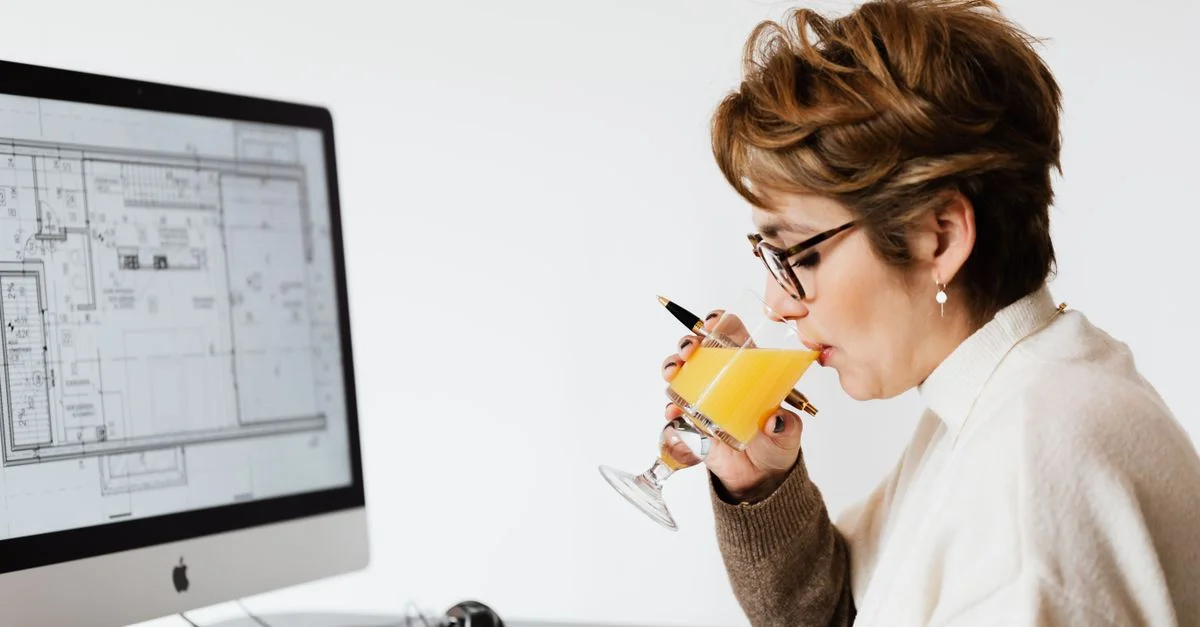 Femme architecte travaillant à son bureau, concentrée sur des plans de design informatique, buvant du jus.