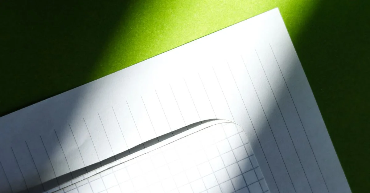Vue en gros plan de feuilles de papier ligné et graphique sous un soleil éclatant projetant des ombres sur une surface verte.