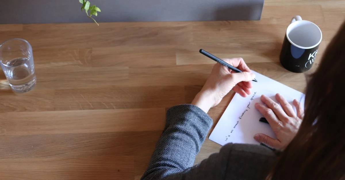 Une femme adulte prenant des notes sur du papier à un bureau en bois dans un cadre de bureau avec une tasse et un verre d'eau.