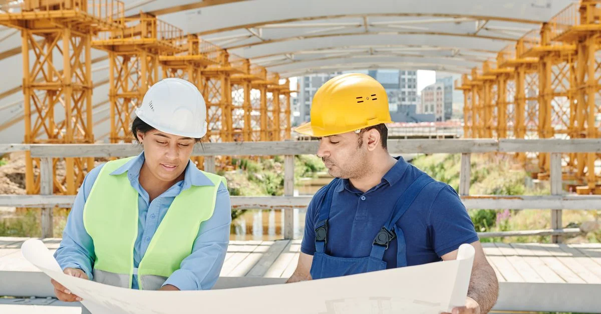 Équipe d'ingénieurs analysant des plans ensemble sur un chantier de construction en extérieur avec des équipements de sécurité.