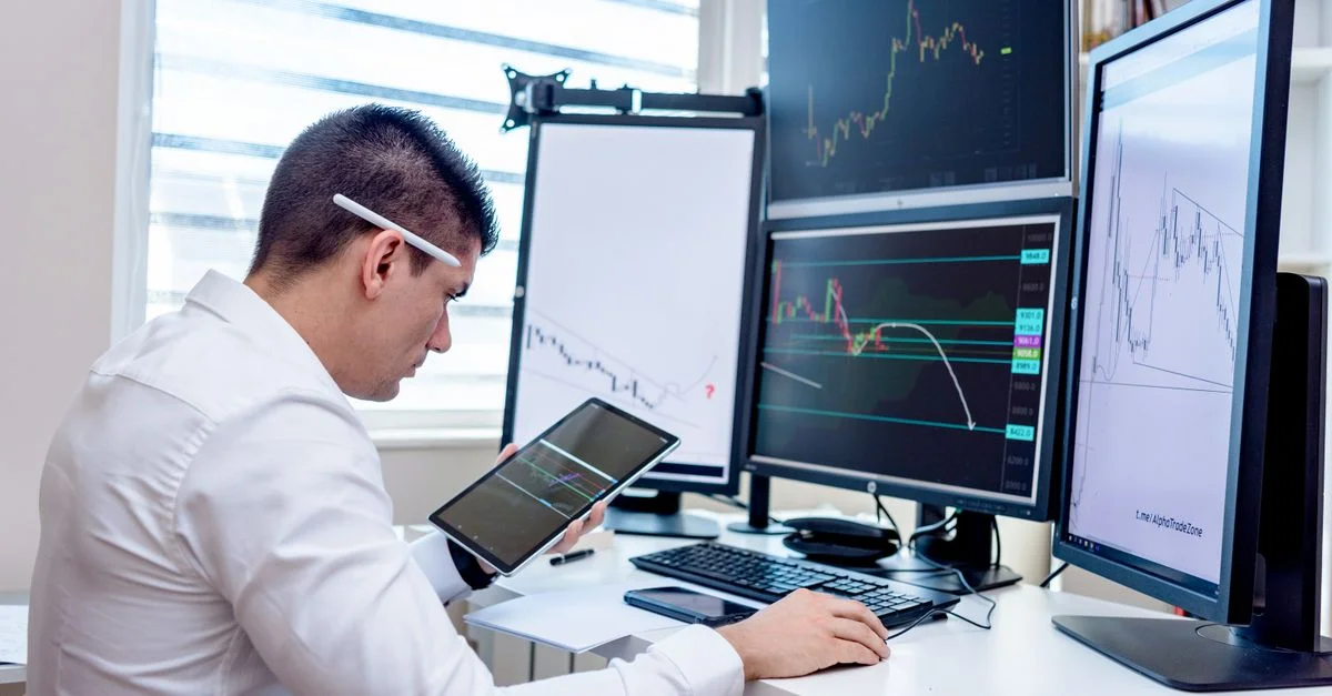 Professionnel analysant des graphiques de marché boursier sur plusieurs moniteurs à son bureau.