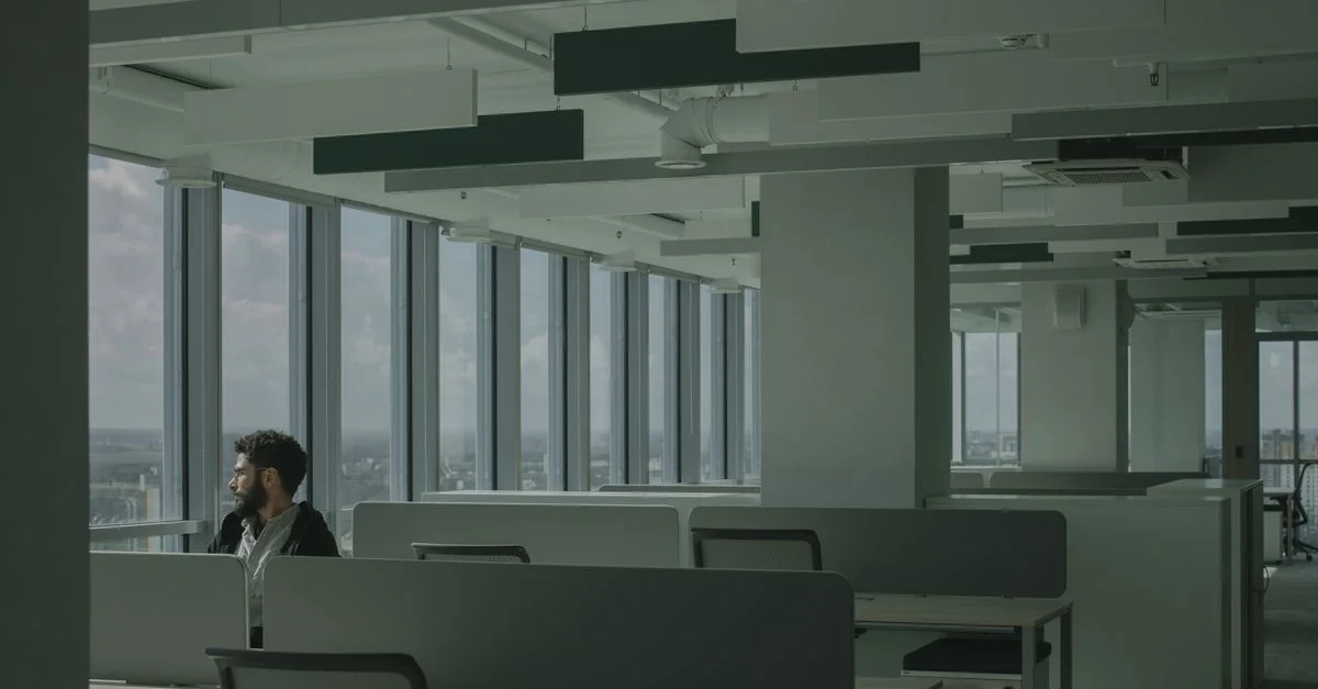 Un homme est assis seul dans un espace de bureau moderne avec de grandes fenêtres donnant sur un paysage urbain.