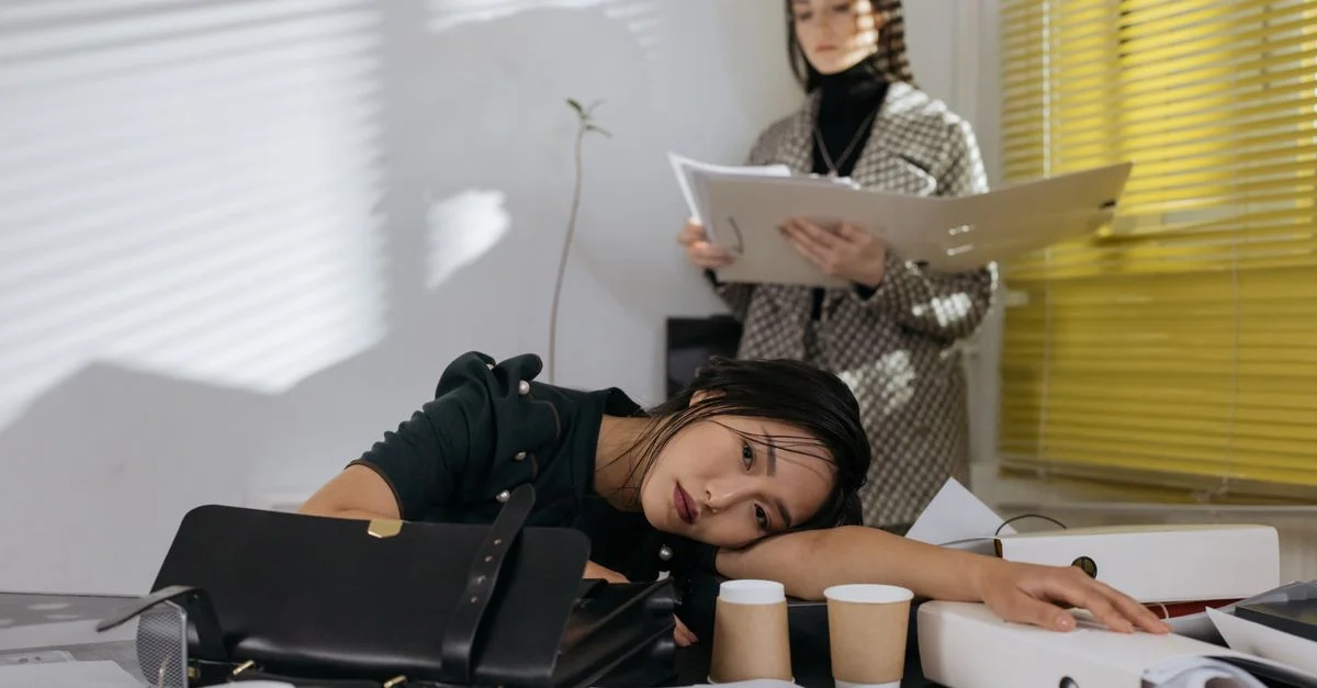 Employée épuisée reposant sur le bureau entourée de documents, collègue en arrière-plan.