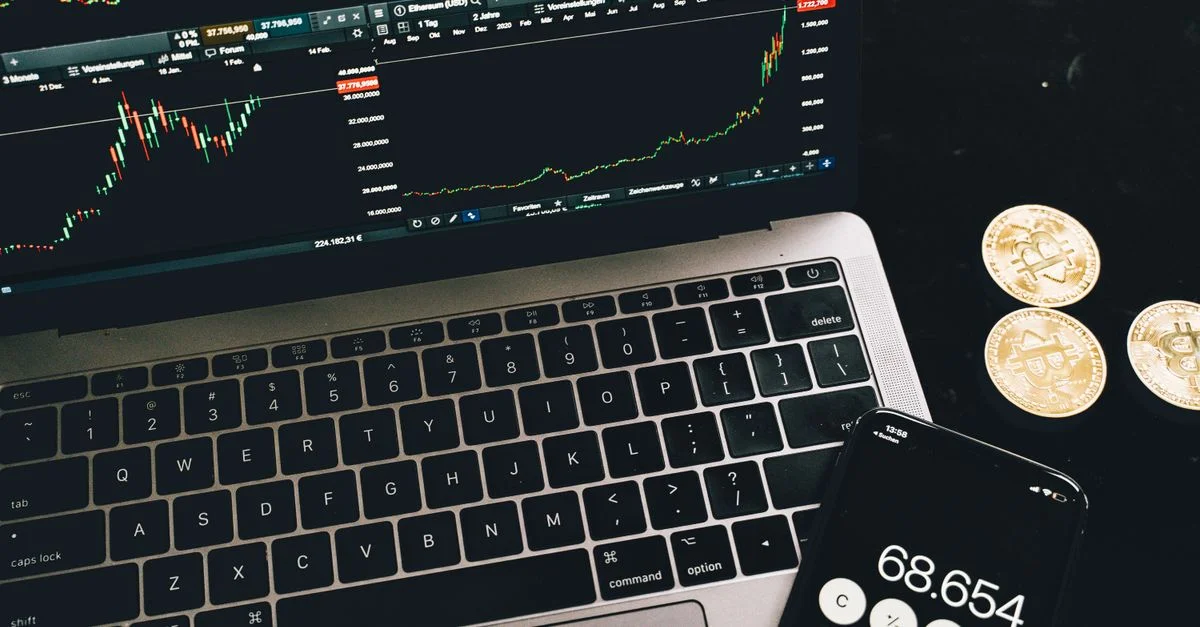 Un ordinateur portable avec des graphiques de trading, une calculatrice smartphone et des pièces de bitcoin représentant le trading de cryptomonnaie.