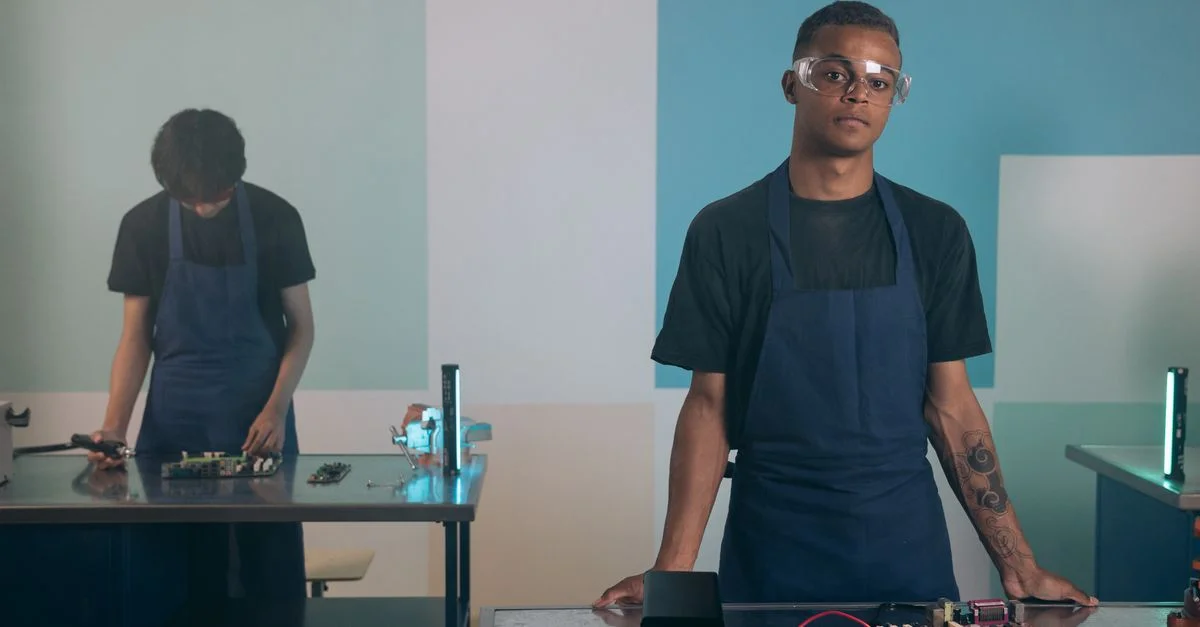 Deux hommes travaillant sur des appareils électroniques dans un atelier, portant des tabliers et des lunettes de protection.