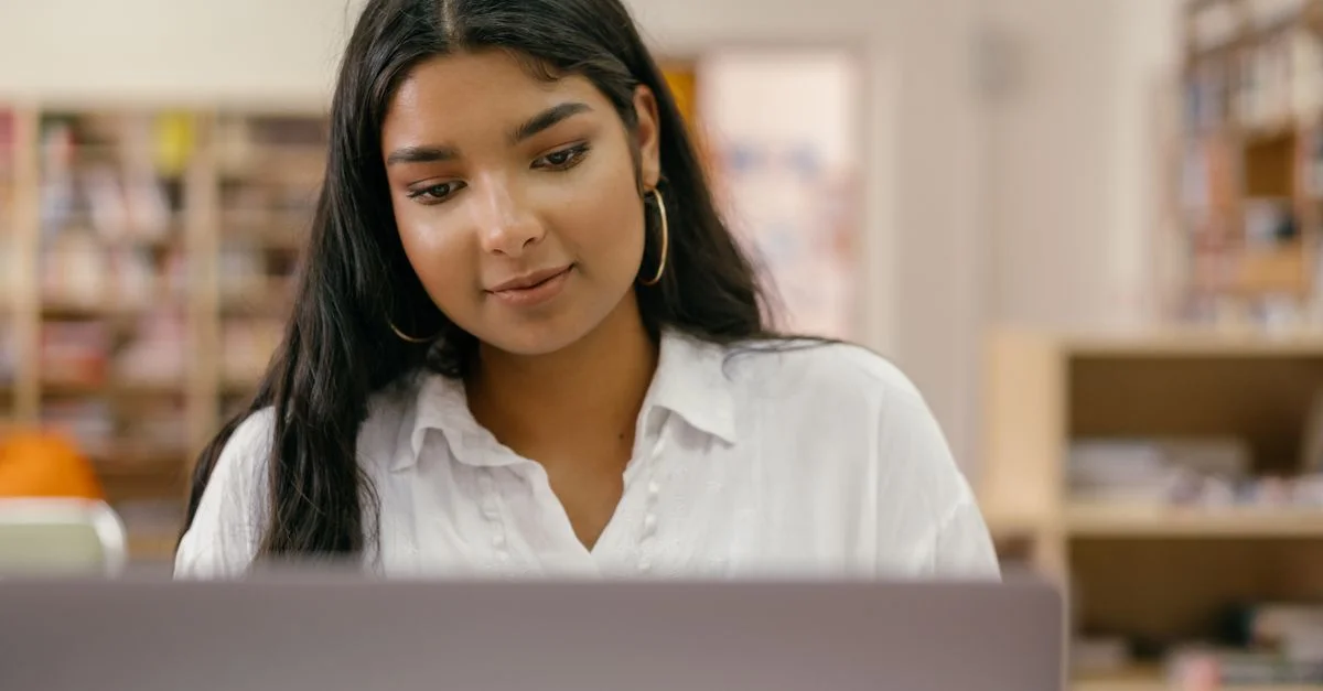 Une femme pensive utilisant un ordinateur portable dans un cadre de bibliothèque, concentrée sur son travail.