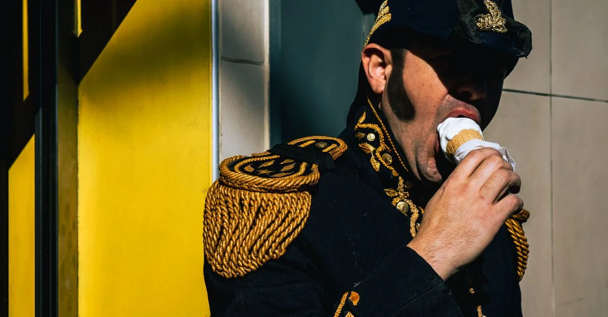 Un homme vêtu d'un uniforme militaire historique mange une glace contre un mur jaune.