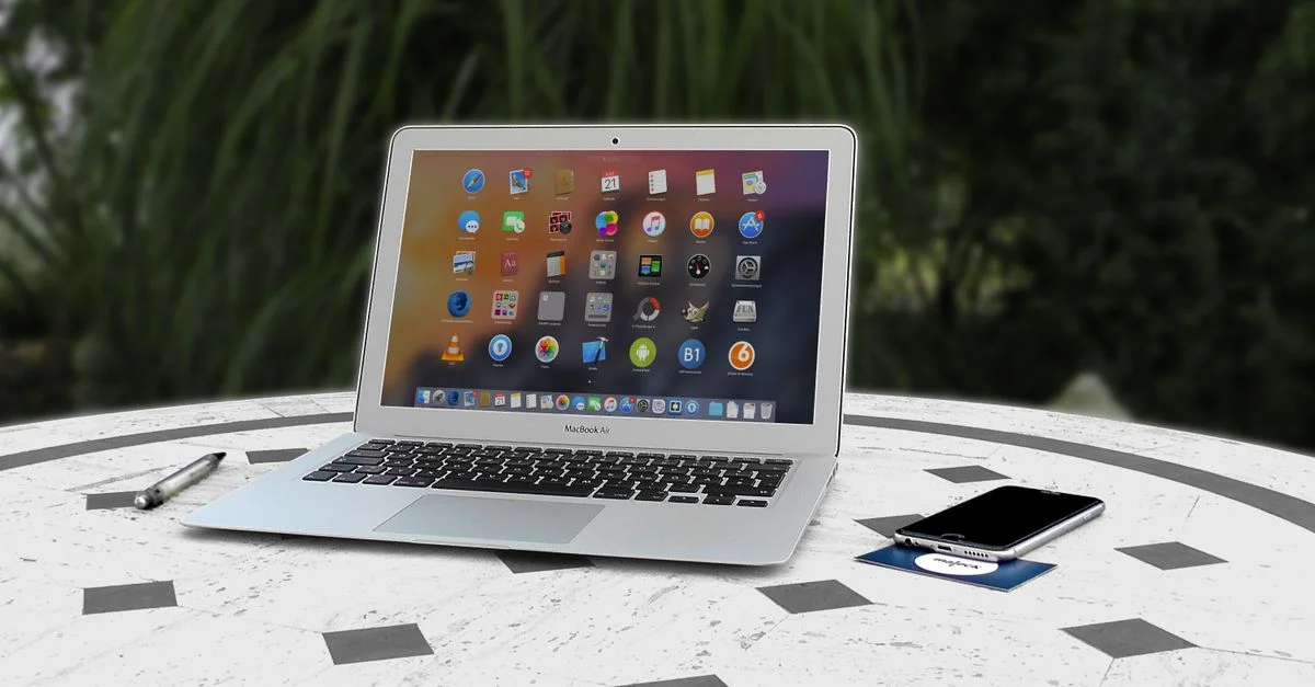 Ordinateur portable ouvert avec des icônes d'applications affichées, un smartphone et un stylo sur une table blanche en extérieur.