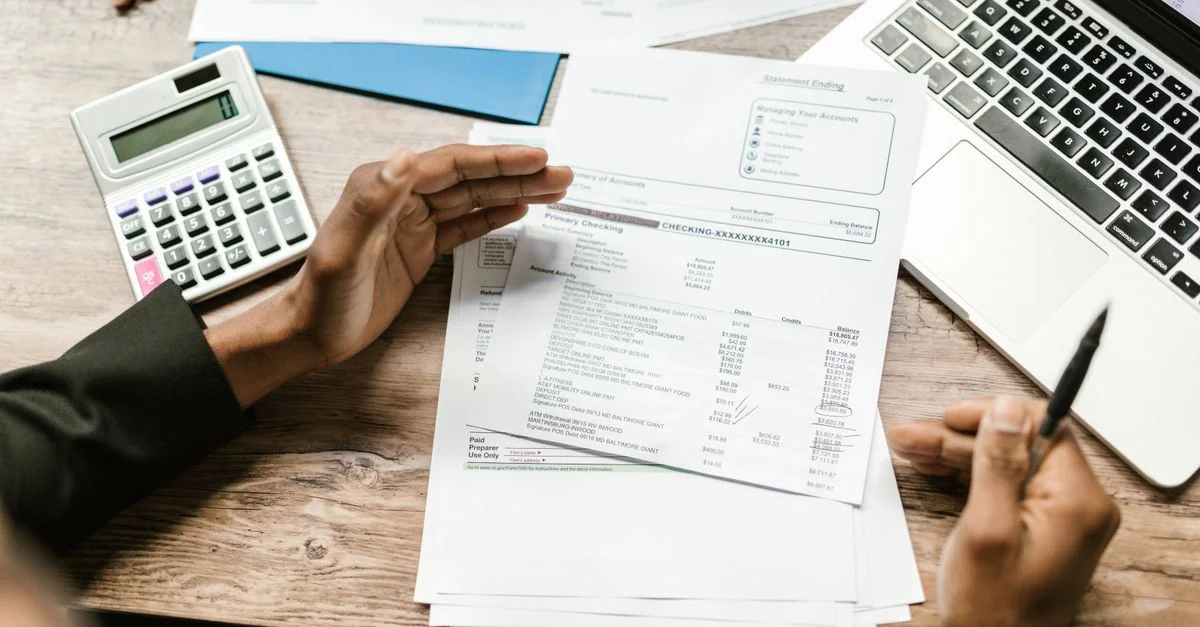 Mains tenant des documents financiers avec une calculatrice et un ordinateur portable sur un bureau, scène d'analyse commerciale.