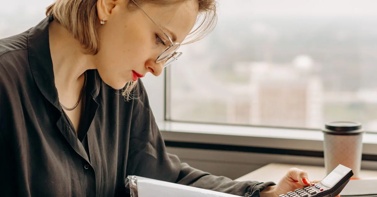 Femme d'affaires travaillant avec une calculatrice et des documents financiers près d'une fenêtre, concentrée et analytique.