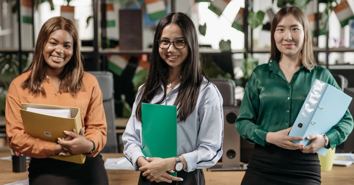 Trois femmes diverses souriantes tenant des dossiers dans un environnement de bureau moderne.