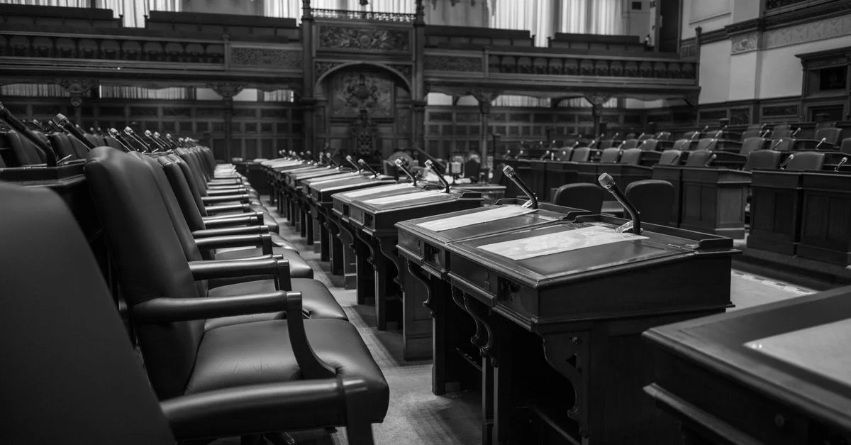 Photographie en noir et blanc d'une salle d'assemblée législative traditionnelle, avec des sièges et des bureaux vides.