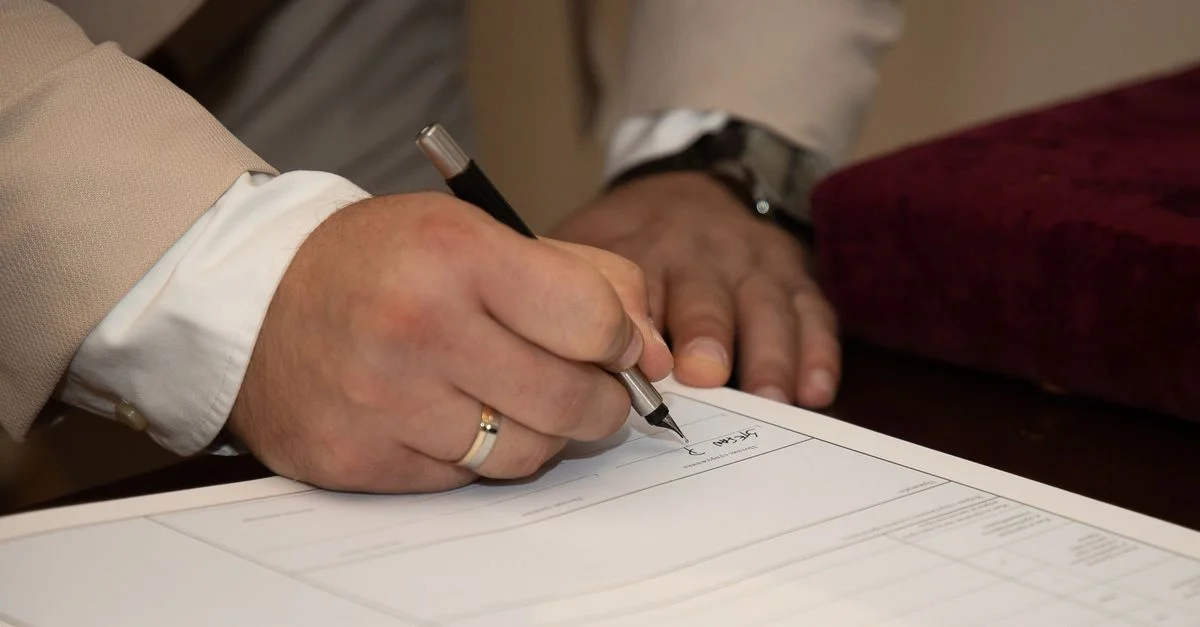 Gros plan d'un homme signant un document, mettant en valeur une alliance et un stylo.