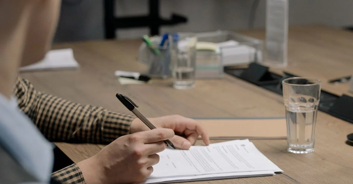 Personne en tenue professionnelle signant un document sur une table en bois dans un cadre de bureau.