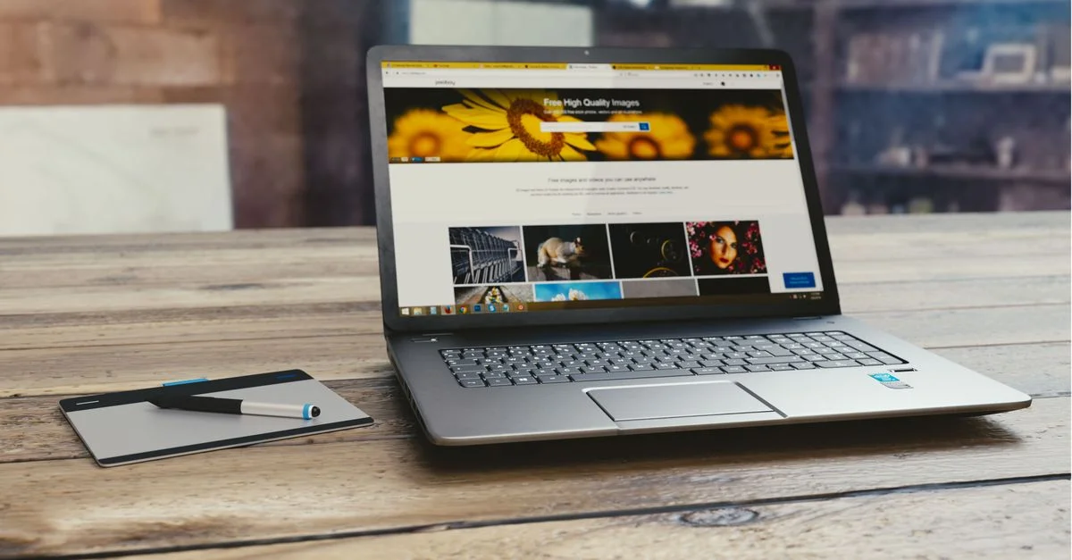 Ordinateur portable avec page web ouverte sur bureau en bois, accompagné d'une tablette et d'un stylo, représentant un environnement de travail numérique.