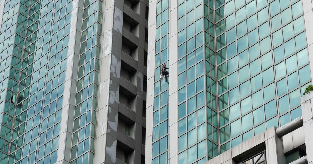 Un travailleur nettoie les vitres d'un haut gratte-ciel à l'aide d'un harnais et de cordes.