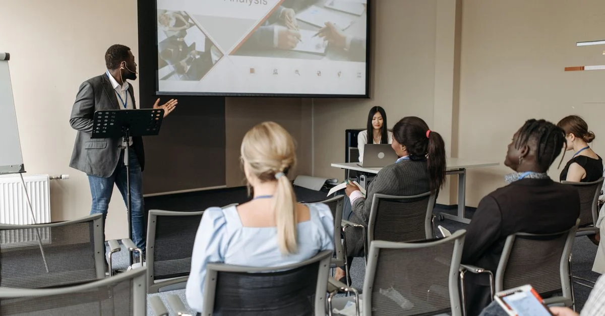 Un groupe divers assiste à un séminaire d'analyse commerciale avec un présentateur et un écran de projection dans une salle de conférence moderne.