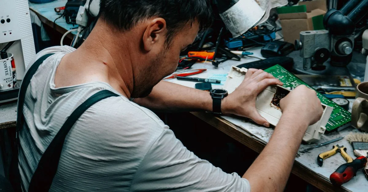 Un homme travaille intensément à la réparation d'appareils électroniques sur un établi d'atelier encombré.