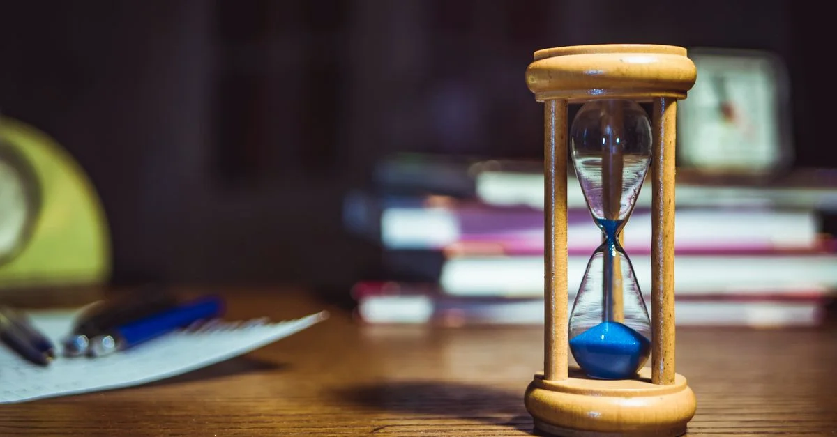 Gros plan sur un sablier en bois sur un bureau, aux côtés de livres et de stylos, suggérant une étude concentrée ou une gestion du temps.