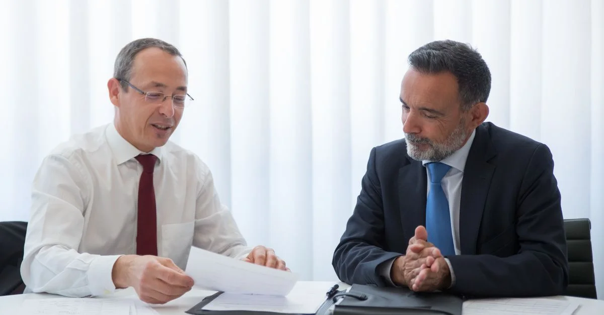 Deux hommes d'affaires en costume discutent de documents dans un bureau lumineux.