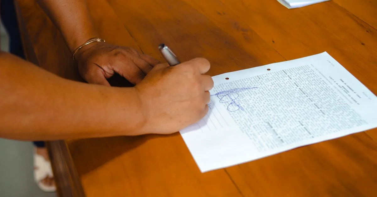 Gros plan de mains signant un document juridique sur une table en bois à l'intérieur.