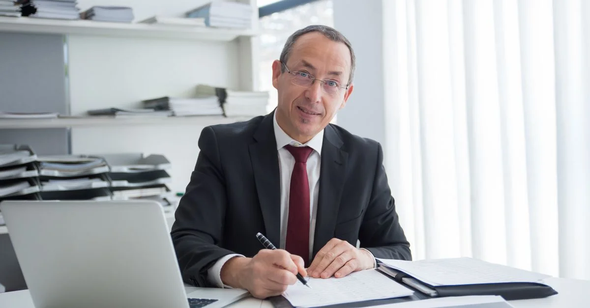 Un homme d'affaires en costume écrivant dans un bureau avec un ordinateur portable et des documents.