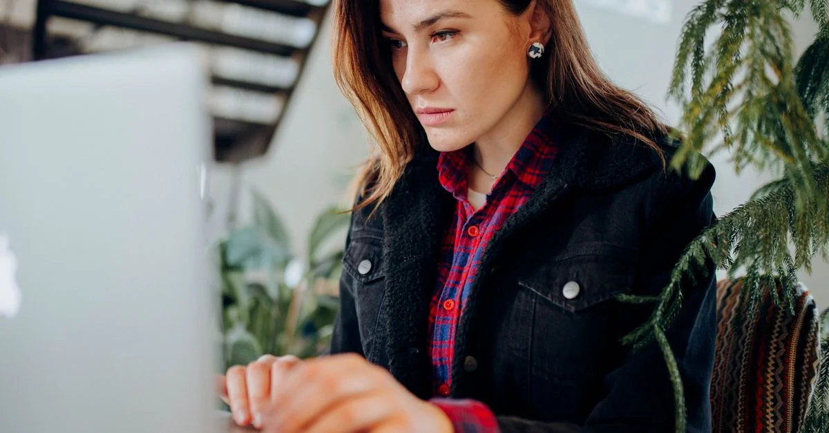 Une femme sérieuse en chemise à carreaux travaille intensément sur son ordinateur portable à l'intérieur.
