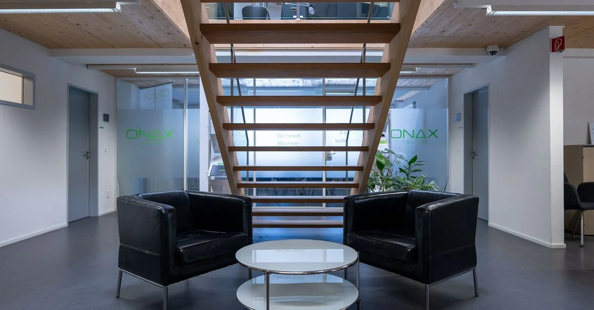 Salon de bureau contemporain à Cazis avec des chaises noires élégantes et une table en verre sous un escalier en bois.
