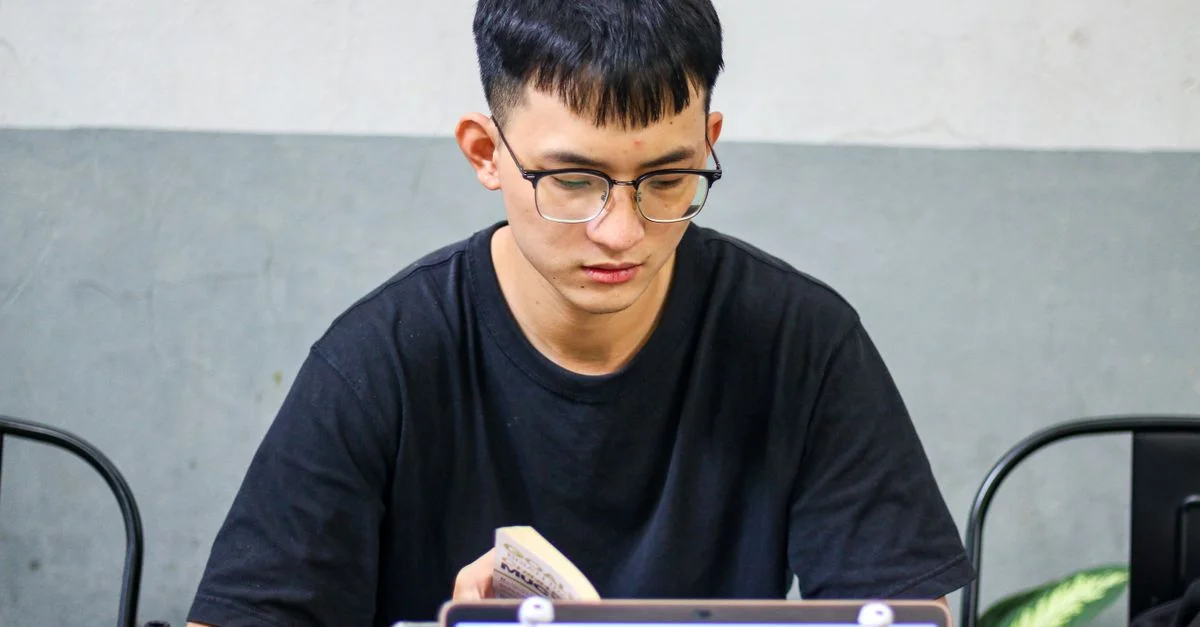 Adolescent portant des lunettes lisant un livre à un bureau intérieur avec un ordinateur portable.