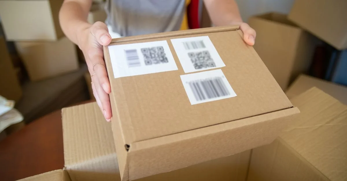 Gros plan sur une personne tenant une boîte en carton avec des étiquettes de code-barres, dans un cadre intérieur.