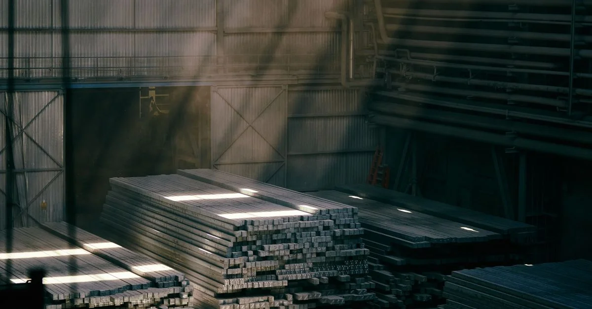 La lumière du soleil pénètre dans un entrepôt industriel et illumine des poutres d'acier empilées.
