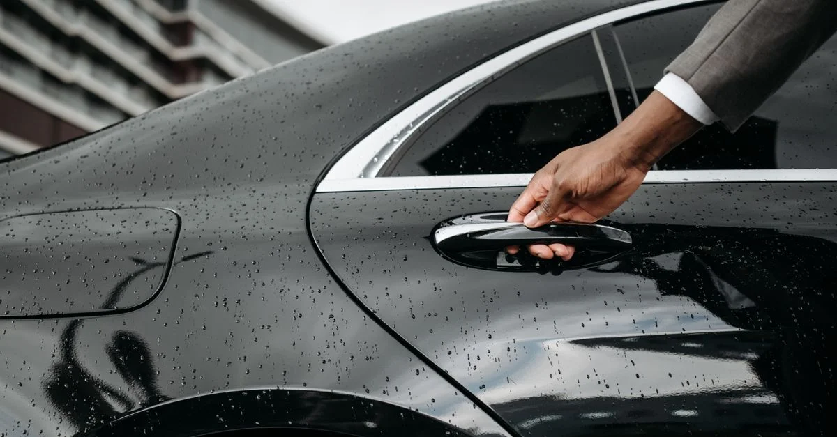 Un gros plan d'une main ouvrant une porte de voiture noire couverte de gouttes de pluie, mettant en valeur l'élégance et les détails météorologiques.