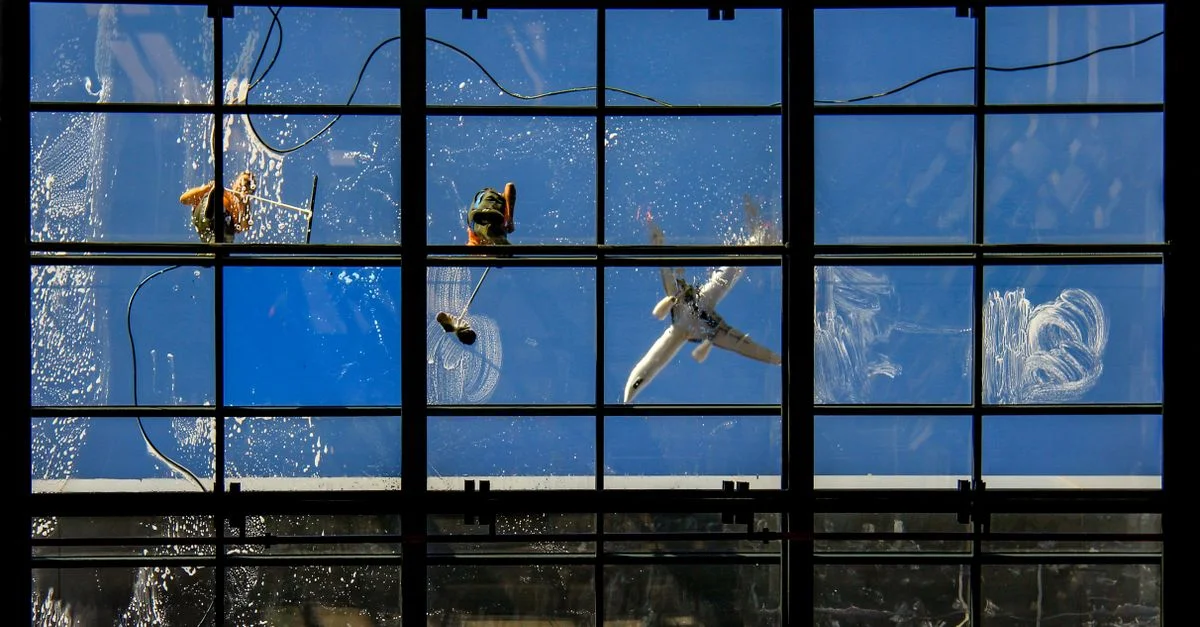 Des laveurs de vitres au travail, un avion survolant la zone sur fond de ciel bleu.