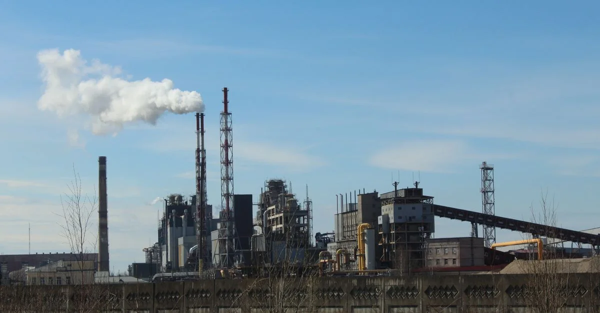 Photo capturant une usine industrielle avec des cheminées émettant de la fumée sous un ciel bleu clair.