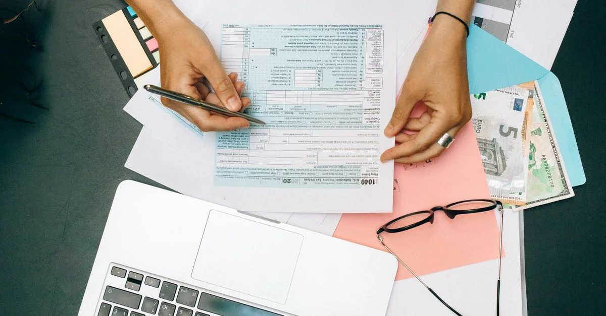 Mains écrivant sur des documents fiscaux avec un ordinateur portable, des lunettes et de la monnaie sur le bureau.