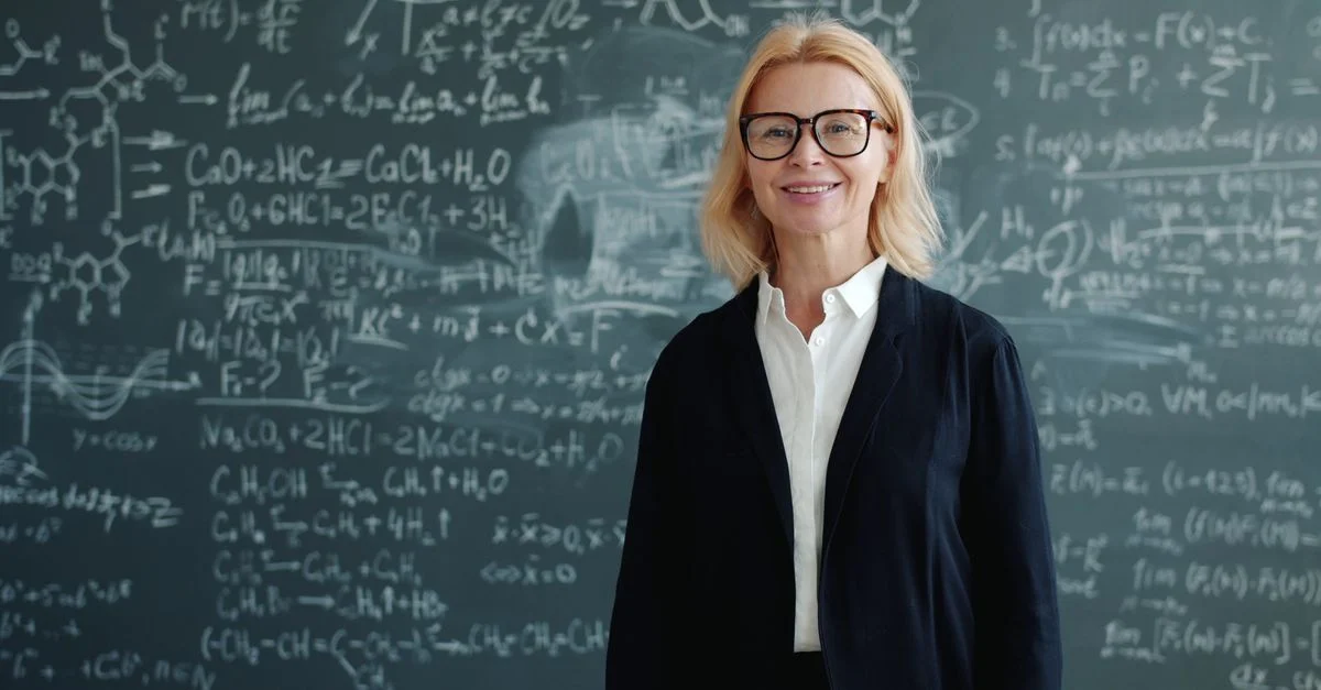 Une enseignante souriante devant un tableau noir couvert d'équations complexes.