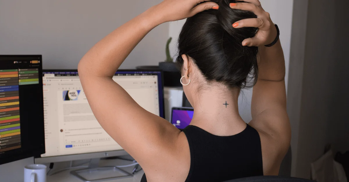 Une femme avec un tatouage relève ses cheveux dans un espace de travail moderne avec des écrans doubles.